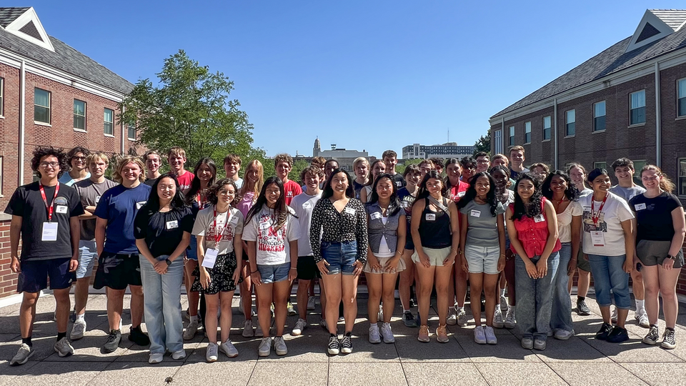 Group of young adults posing outdoors, surrounded by brick buildings under a bright blue sky.