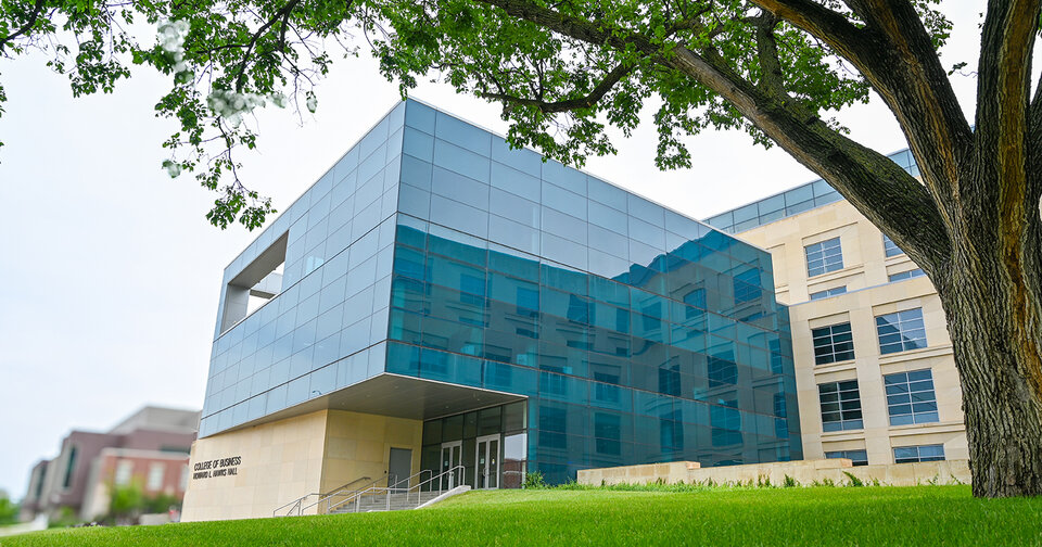 A modern building with large blue-tinted glass windows and beige stone walls, surrounded by green grass and a tree in the foreground.