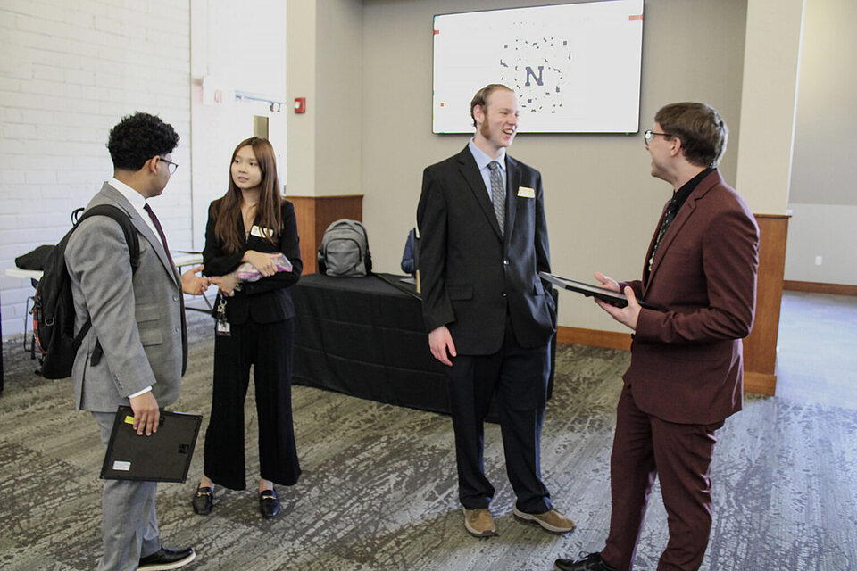 Group of people in formal attire talking in a room with a display screen and table in the background.