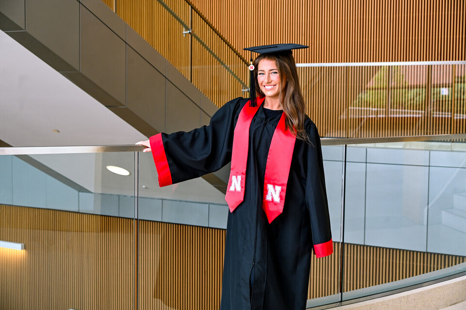 Graduate in black gown with red accents standing near a glass railing indoors.