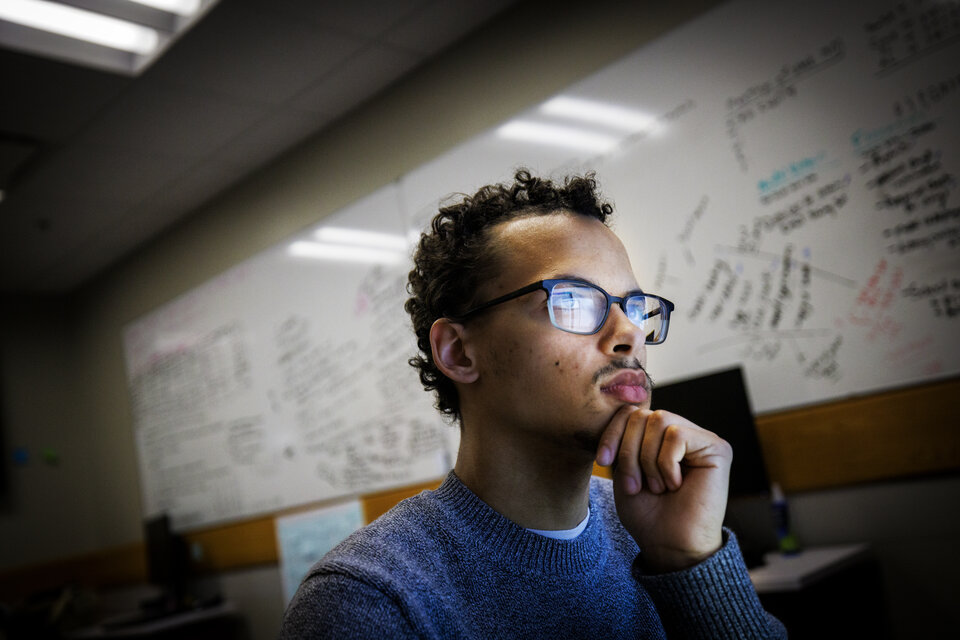 One student looking at their computer intently. The photo is taken facing the student with a whiteboard in the background containing writing..