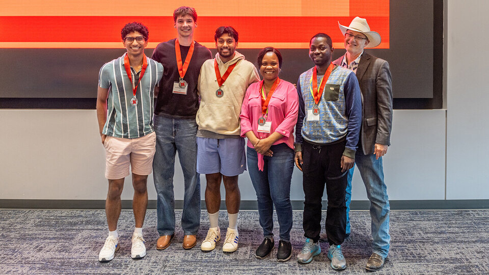 Group of six people wearing medals, standing indoors in front of a screen.