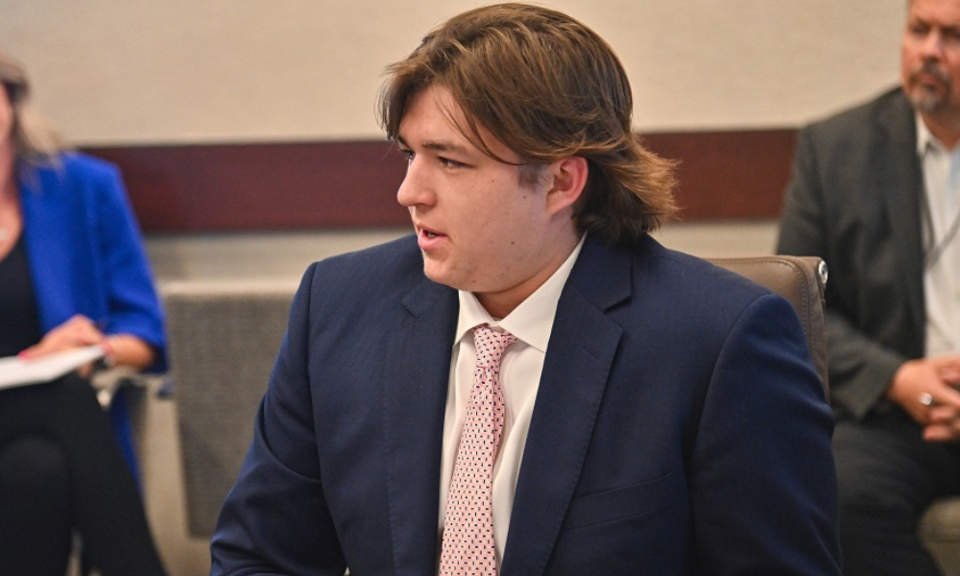 A picture of a man wearing a navy blue suit jacket, a white shirt, and a pink tie, with medium length hair, sat at a table and talking.