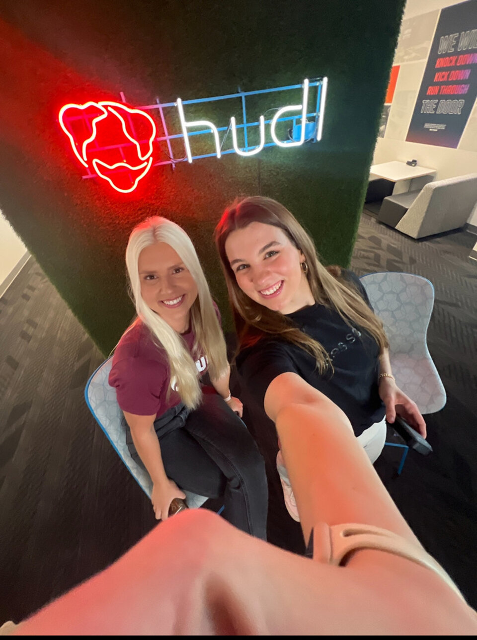 Two people sat next to one another on chairs in front of a large neon sign, smiling and taking a selfie together.