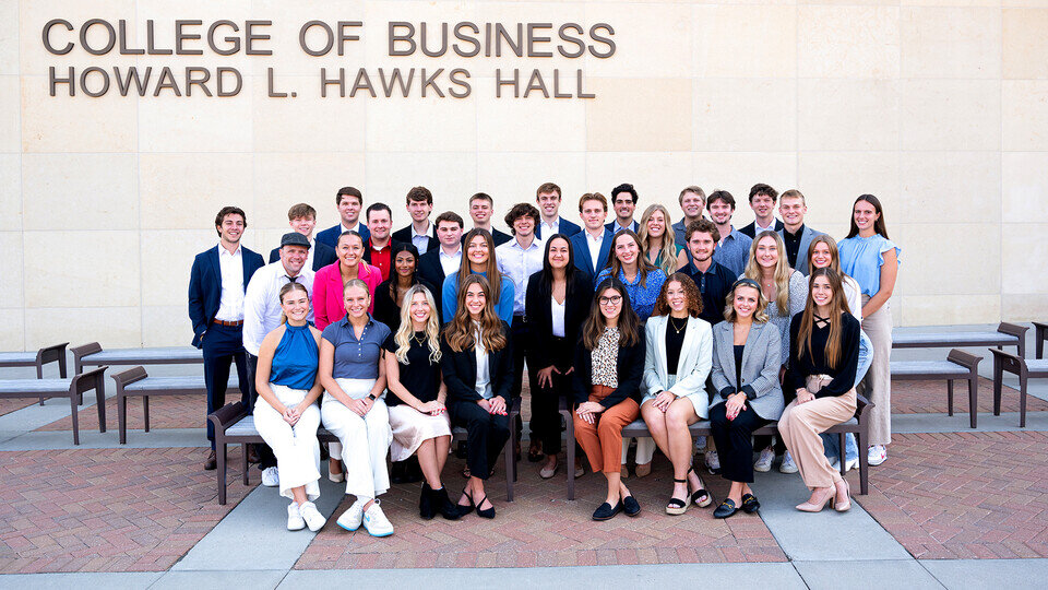 A group of people assembled for a group portrait on a red brick footpath in front of a beige stone building with the text "College of Business, Howard L. Hawks Hall".