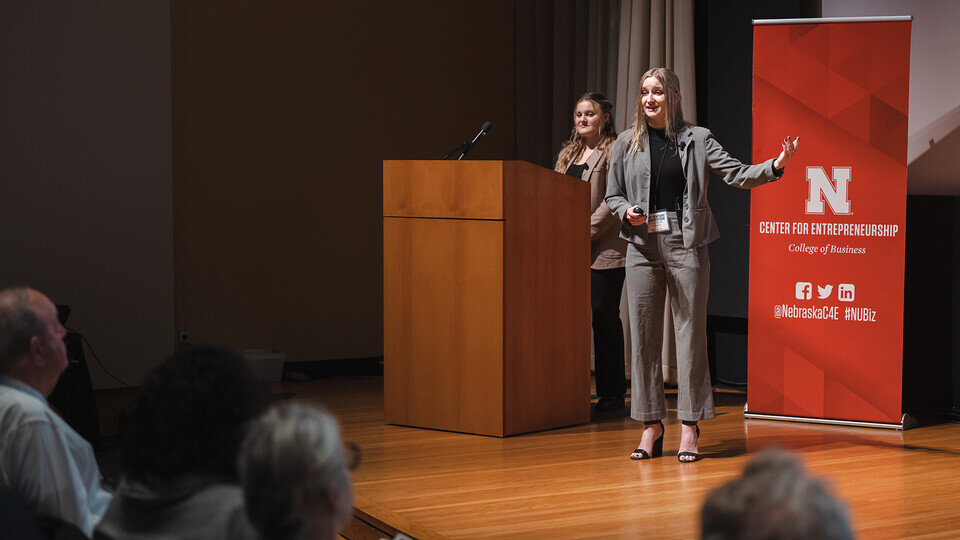 Two people standing on a stage next to a podium and a banner sign, presenting towards an audience in the foreground.