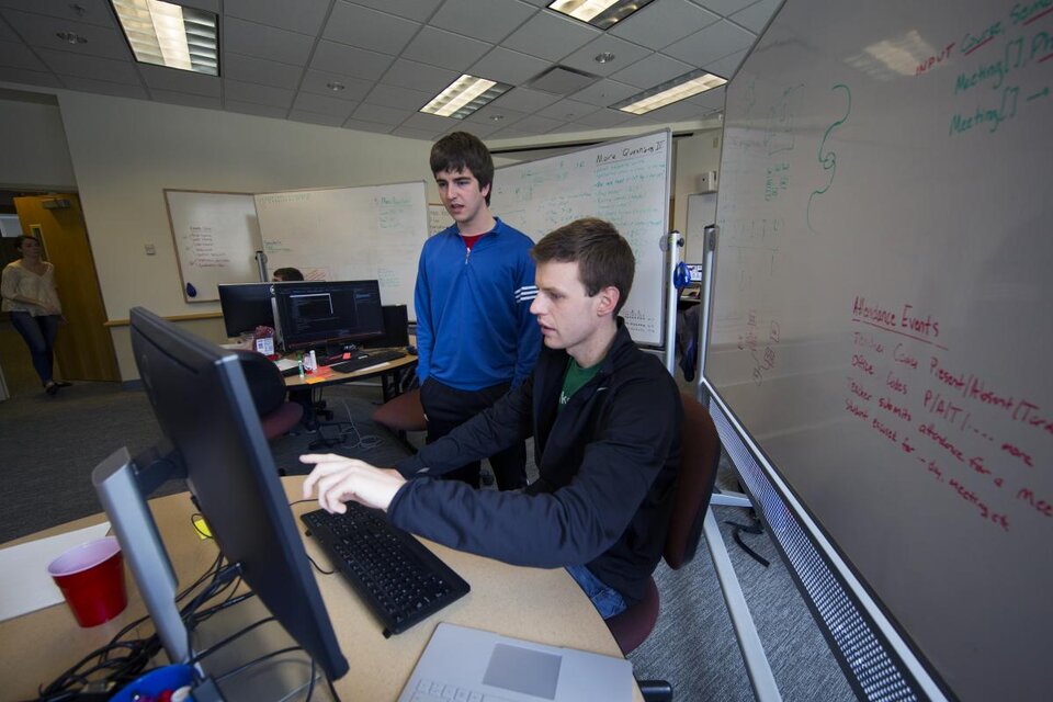 A person sat in a char pointing at a computer monitor whilst a person stands next to them looking at the monitor, with dry erase boards in the background.