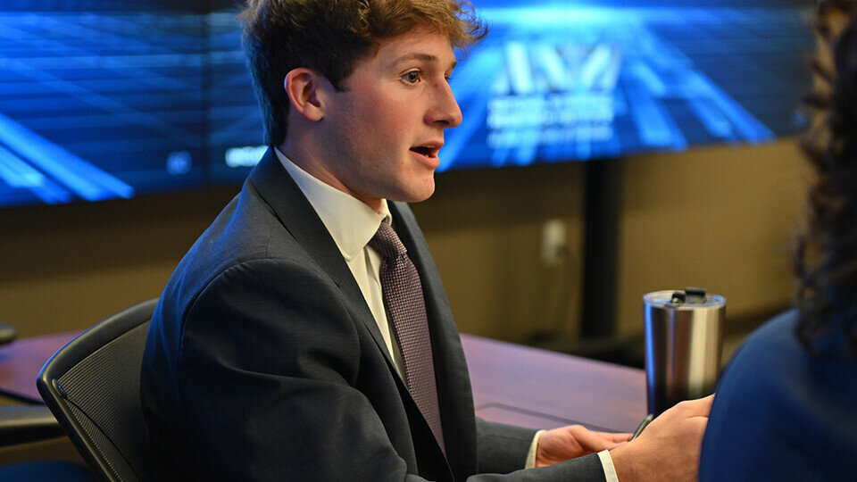 A man wearing a dark grey suit, a white shirt, and a brown tie, sat at a table and talking.