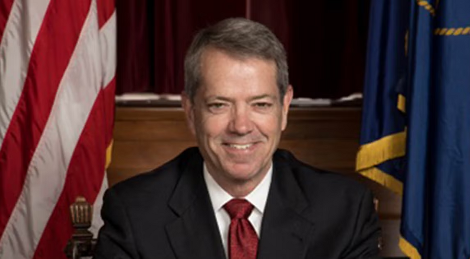 A man wearing a black suit with a red tie and white shirt sits in front of two flags and smiles at the camera.