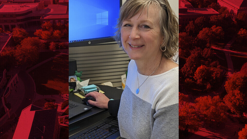 A person in a white and grey sweater sat at a computer holding a mouse and smiling at the camera.