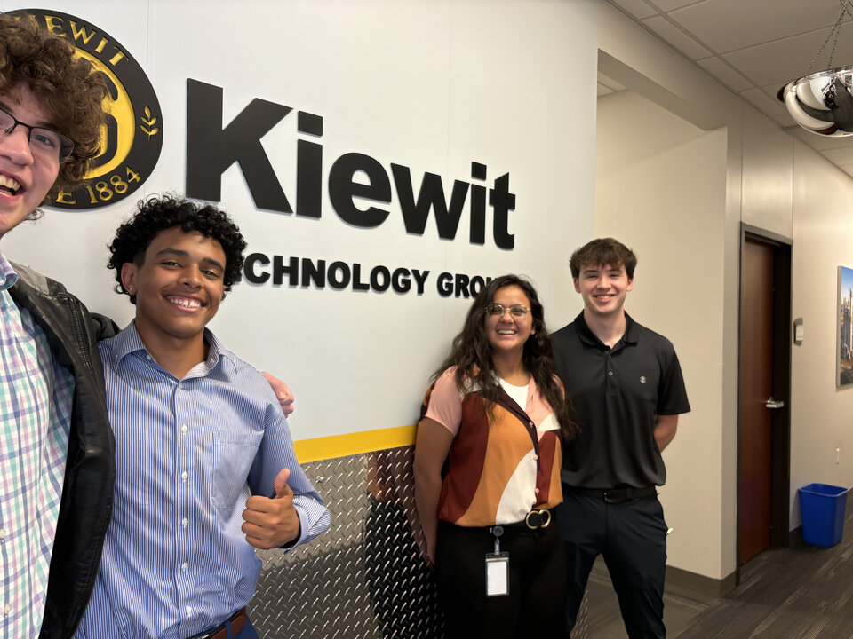 Four students standing in front of the Kiewit Technology Group wall sign