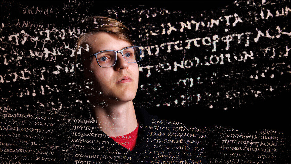 A picture of a man wearing glasses and a black t-shirt looks at a variety of letters floating in front of him.