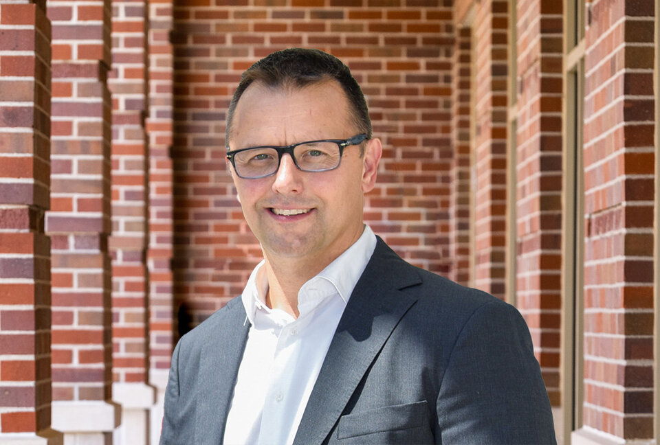 A photograph of a person in a grey blazer and white shirt stood in front of a brick wall.