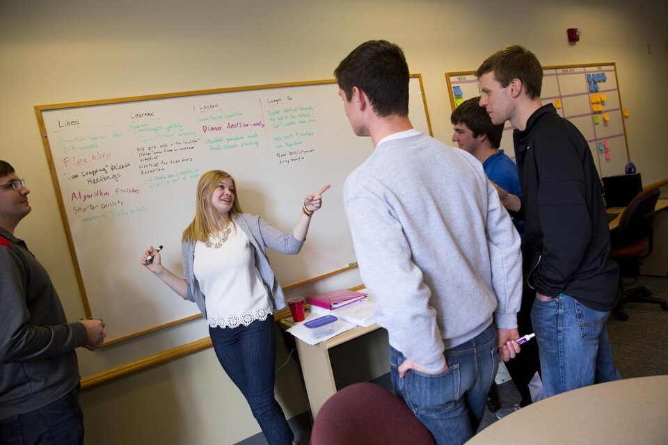 A person stood in front of a dry erase board, pointing at an audience of other people who are looking towards the dry erase board.