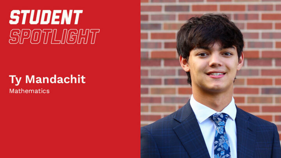 A student with short black hair stands against a brick wall whilst wearing a blue suit and a floral tie.