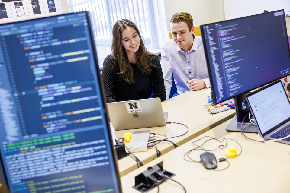 two students looking at computer screen smiling with two monitors displaying code in the forefront